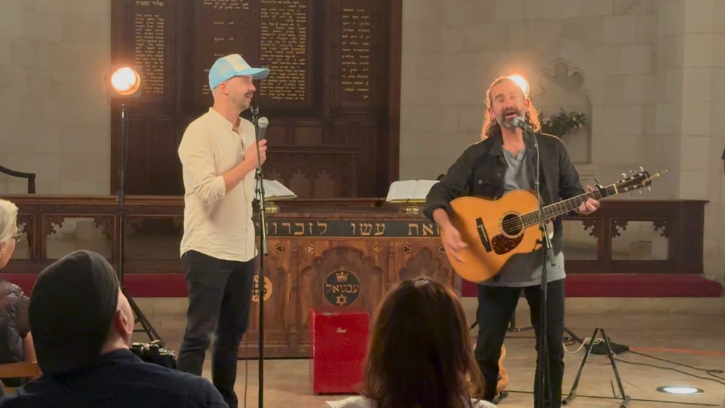 Two musicians performing with a guitar and microphone in a church setting, with a backdrop of a beautifully decorated wall featuring Hebrew text.