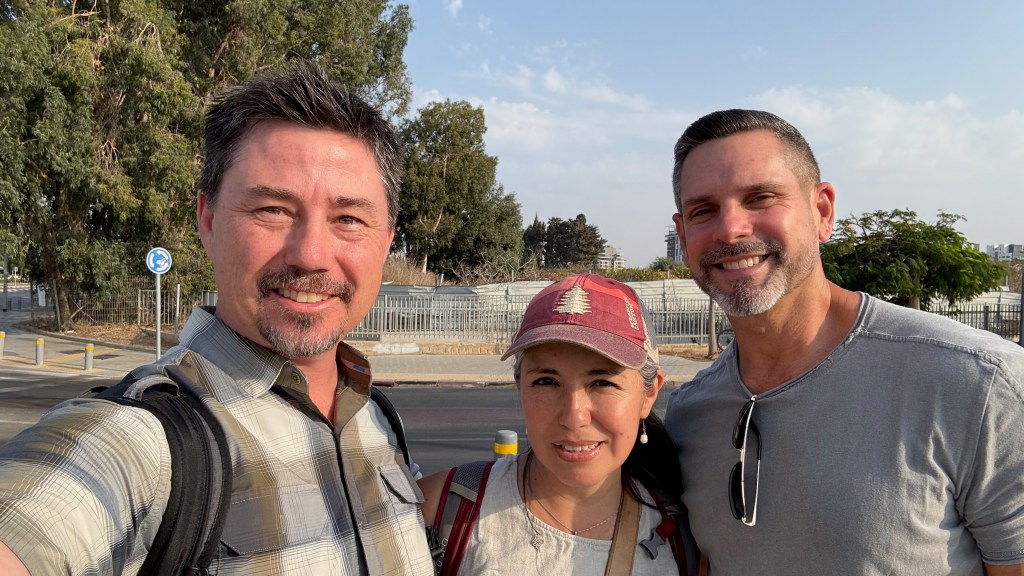 A group selfie featuring three people standing outdoors, smiling at the camera with trees and buildings in the background.