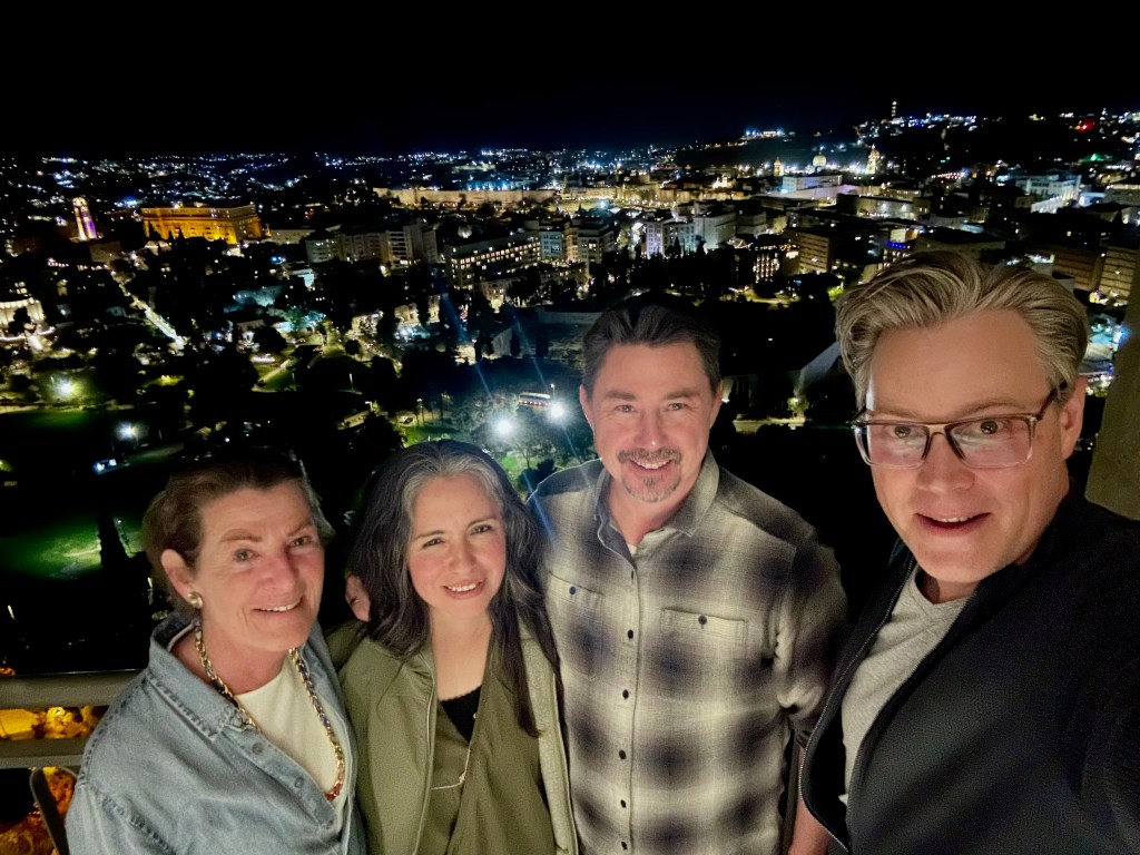 A group of four people posing for a selfie with a cityscape view at night, showcasing illuminated buildings and streets in the background.