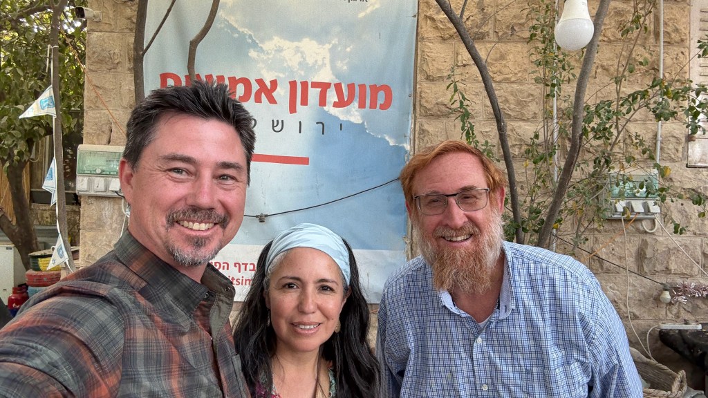 A group selfie featuring three individuals smiling in front of a stone wall with a blurred sign in the background. The scene is set outdoors, capturing a warm and friendly moment.